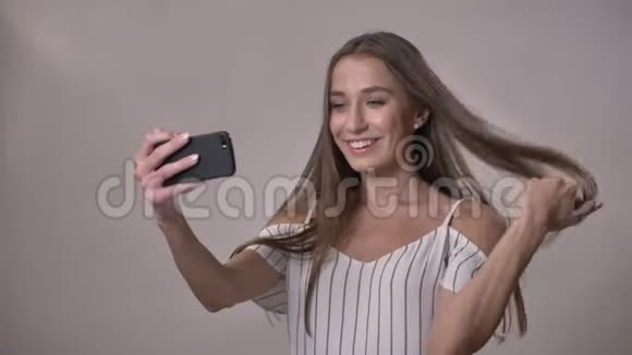 年轻迷人的害羞女孩正在智能手机上制作视频修头发微笑交流理念灰色背景视频的预览图