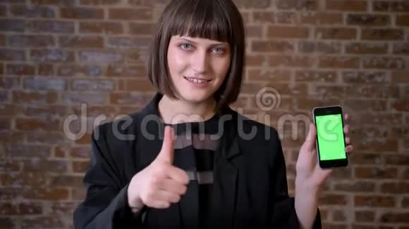 年轻快乐的女人拿着带有色度的电话竖起大拇指砖墙背景视频的预览图