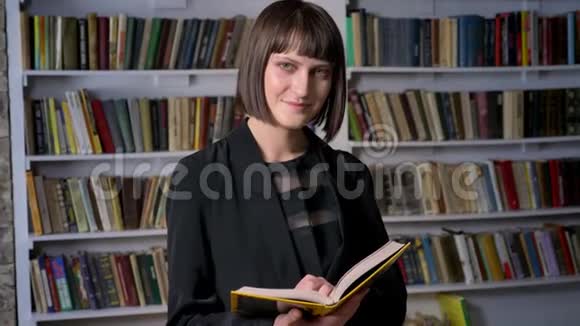 年轻漂亮的女人穿着夹克拿着书在图书馆里看着相机微笑着视频的预览图