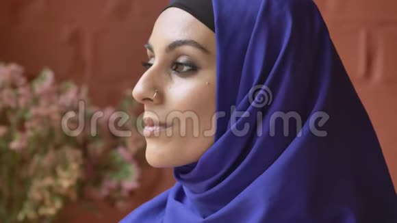 年轻美丽的穆斯林妇女戴着头巾转身看着镜头鲜花和背景墙视频的预览图