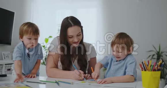 一位年轻的母亲带着两个孩子坐在白色的桌子旁慢慢地在纸上画着彩色铅笔视频的预览图