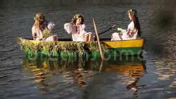 三个女孩穿着斯拉夫民族服装坐着一艘船漂浮在河上穿着花圈的女孩们欢快地摆着姿势和欢笑视频的预览图