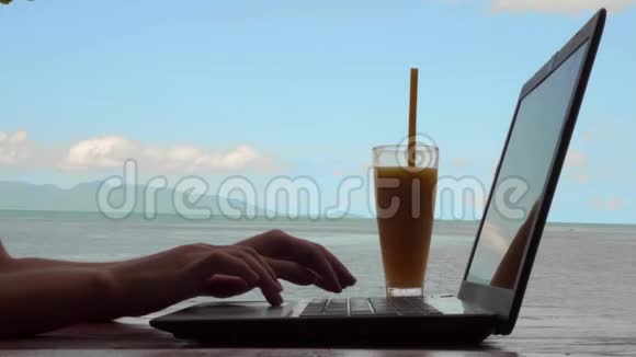 年轻白种人女士使用笔记本电脑和饮用芒果水果奶昔与海景的循环电影远程工作和视频的预览图