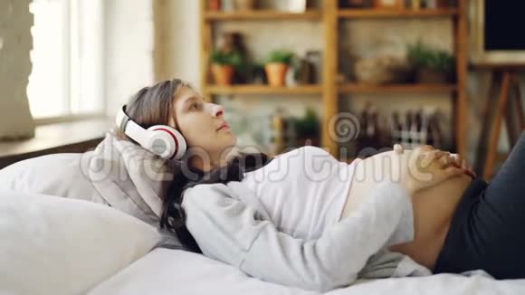 期待着妈妈抚摸着她的肚子听着音乐带着耳机在家放松躺在卧室的床上视频的预览图