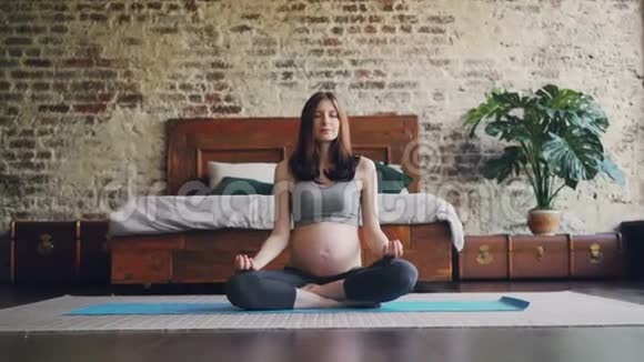 怀孕的女孩坐在瑜伽垫上双手放在膝盖上呼吸和放松享受着平静视频的预览图