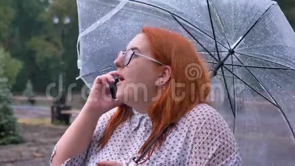带眼镜的胖姜女站在公园的雨下打电话撑伞交流视频的预览图
