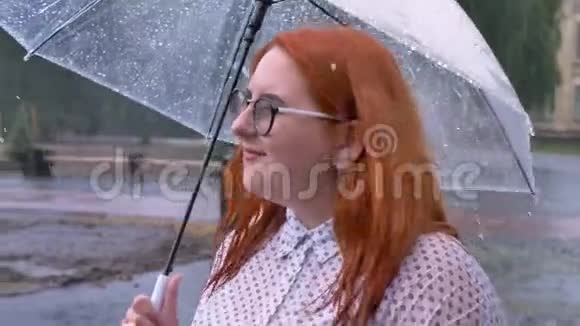 戴着眼镜的肥姜女孩在雨中漫步在公园里看着天空中撑着伞侧目观看视频的预览图