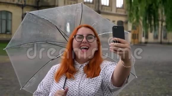 戴着眼镜的肥姜女孩正在公园里的智能手机上在自拍眨眼拿着伞视频的预览图