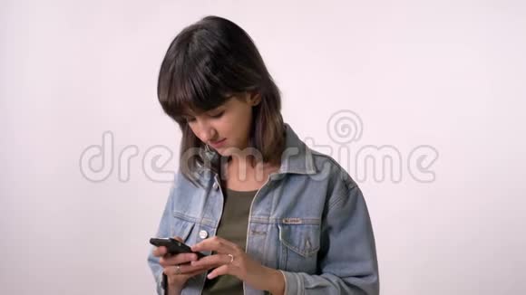 年轻的黑发女孩正在智能手机上输入信息看着摄像机微笑着白色背景视频的预览图