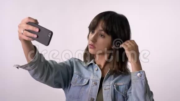 年轻的黑发女孩正在智能手机上自拍鸭脸白色背景视频的预览图