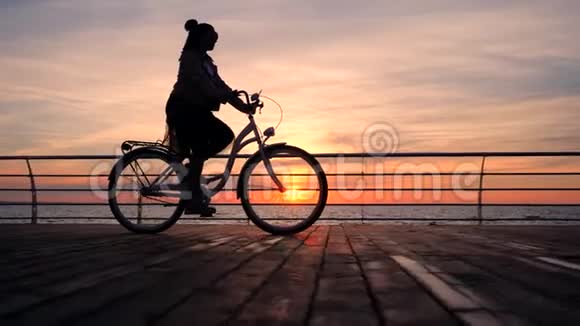 美妙的日出或日落在海洋之上年轻时尚女孩骑着老式自行车在木堤上骑车剪影视频的预览图