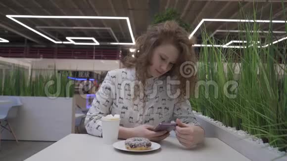 年轻漂亮的红发女人穿着休闲风格坐在咖啡馆里喝着咖啡和甜甜圈视频的预览图