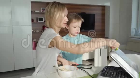 年轻的女人在厨房做华夫饼可爱的男孩帮助她一大早就不规律地做早餐视频的预览图