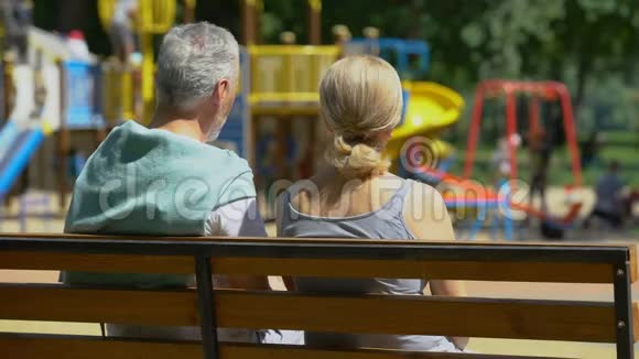退休的男人和女人坐在公园的长椅上看着孙子快乐的回忆视频的预览图