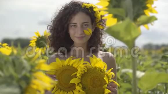 一幅美丽的卷曲嬉戏的女孩看着镜头微笑地站在向日葵田野的肖像明亮的黄色视频的预览图