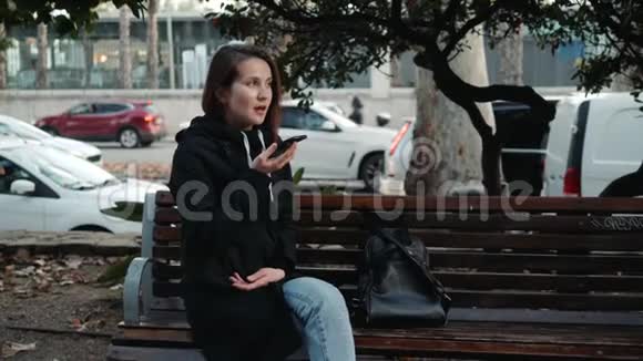 一位微笑的时髦女孩坐在公园的长椅上用手机打字留言的肖像音频语音视频的预览图