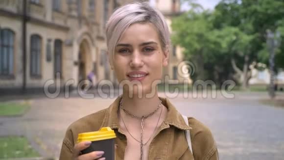 金发快乐的年轻女子鼻子穿孔拿着咖啡看着相机站在城市的街道上视频的预览图