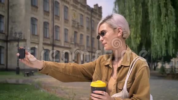 戴着太阳镜的年轻金发女子站在大学附近的公园里自拍并端着咖啡视频的预览图