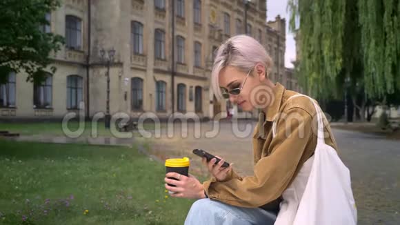 金发美女戴墨镜拿着咖啡打着电话看着相机坐在公园附近视频的预览图