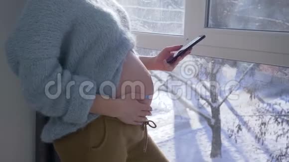 爸爸妈妈怀着宝宝怀孕的女孩站在大窗户旁和男人一起用手机视频的预览图