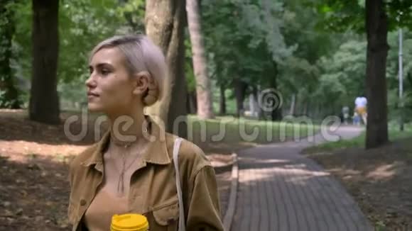 年轻的金发女郎留着短发型在公园散步喝咖啡视频的预览图