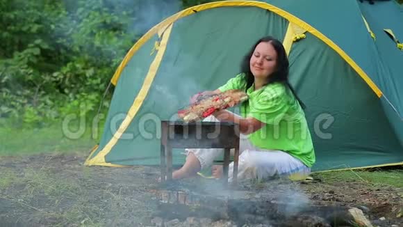 一个年轻的女人坐在一个帐篷背景的森林里在她的烧烤上翻着烤肉视频的预览图