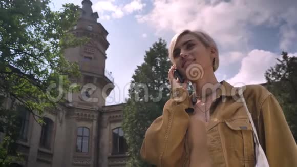 金发迷人的女人短发打电话走开站在大学附近的公园里视频的预览图