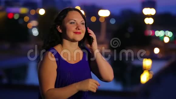 一位穿着蓝色连衣裙的年轻女子正在智能手机上交谈背景是路边模糊的路灯视频的预览图