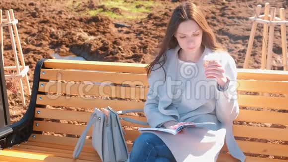年轻漂亮的黑发女子学生坐在春天公园的长凳上看书喝咖啡视频的预览图