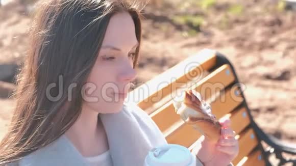 年轻的黑发女人穿着外套坐在城市公园的长凳上吃着糖的面包喝着咖啡春天特写视频的预览图