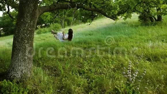 在温暖的季节里留着长发的女孩穿着白色的连衣裙在夏天的橡树下的秋千上摇摆慢动作视频的预览图