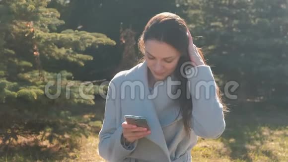 年轻漂亮的黑发女人坐在春天公园的草地上用手机输入一条信息看着相机视频的预览图