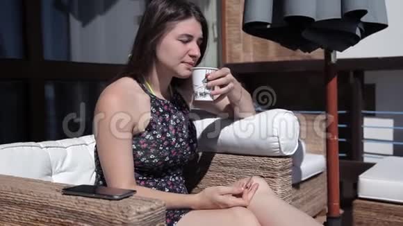 清晨一个年轻的女孩坐在阳台上俯瞰大海喝咖啡视频的预览图