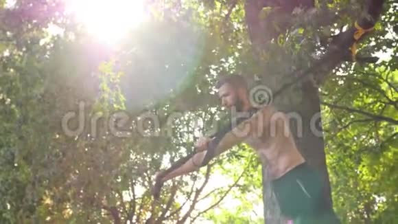 在阳光公园的一棵树旁一个留着胡子的年轻人在健身圈里做俯卧撑4K慢mo镜头闪光效果镜头视频的预览图