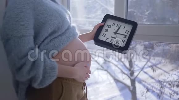 准妈妈站在大窗户旁手里拿着钟抚摸着她赤裸的肚子视频的预览图