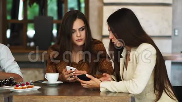 两个女孩坐在咖啡馆里看着电话讨论着她们看到了什么一个人走过来展示了一张有趣的照片视频的预览图