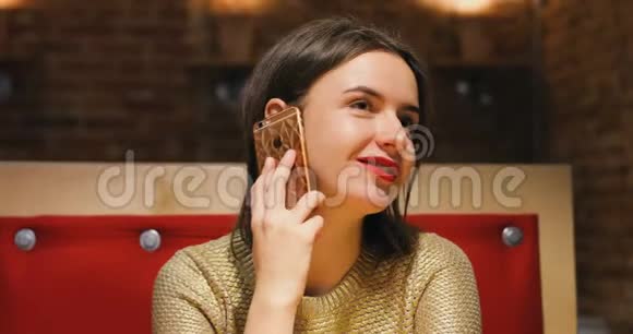 布鲁内特漂亮的年轻女性肖像穿着金色毛衣和红色嘴唇通过智能手机交谈咖啡厅风格背景视频的预览图