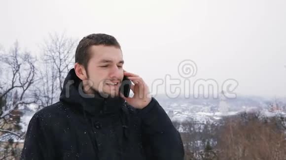 布吕内特年轻男子肖像穿着黑色引擎盖在雪地冬季公园打电话慢动作城市城镇背景视频的预览图