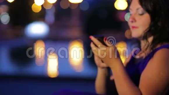 晚上一位穿着蓝色裙子的年轻女性在智能手机上的社交网络中进行交流视频的预览图