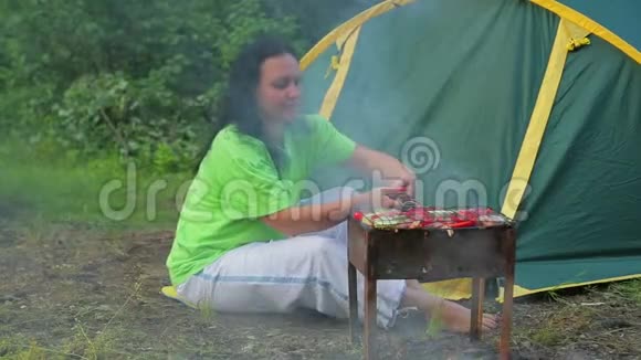 在森林里帐篷背景的年轻女子把烧烤放在烤架上总体计划视频的预览图