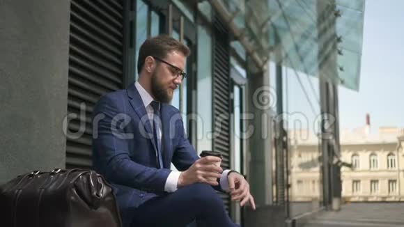 大胡子的商人看着手表坐在城市街道上喝着咖啡视频的预览图