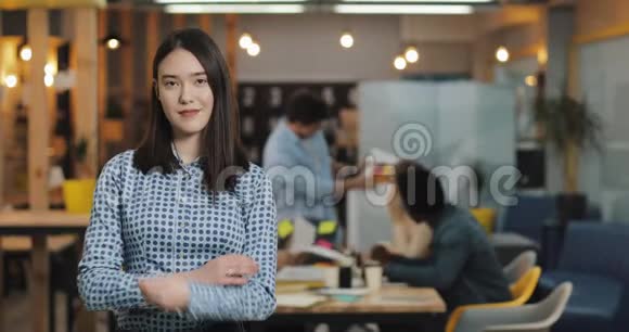 年轻的女商人交叉双臂站在现代的办公室里在模糊的背景下工作的人视频的预览图