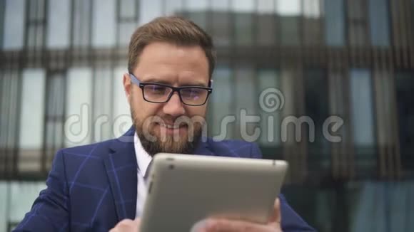年轻的胡子企业主正在使用平板电脑在办公楼背景上微笑视频的预览图