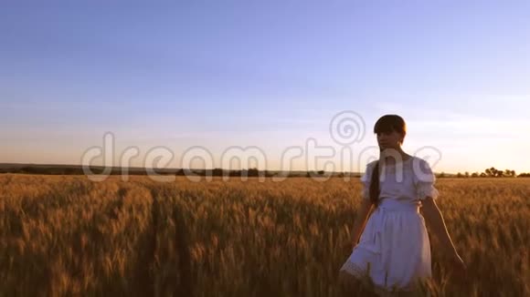 美丽的女孩走过田野带着金色的小麦在夕阳的光辉和微笑中慢动作视频的预览图