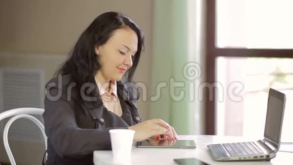一个年轻的女人坐在一家有笔记本电脑和平板电脑的咖啡馆和平板电脑浏览网站慢节奏视频的预览图