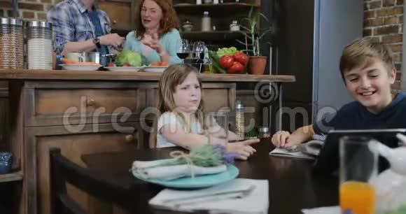 两个孩子坐在厨房的桌子上看滑稽视频而母亲和父亲一起做饭幸福的家庭在家里视频的预览图