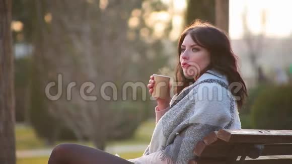 侧景特写迷人可爱的黑发女孩坐在城市公园的长凳上喝热咖啡其中有蒸汽视频的预览图