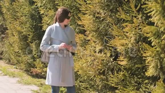 戴太阳镜和外套的年轻女人在城市公园等人春天视频的预览图