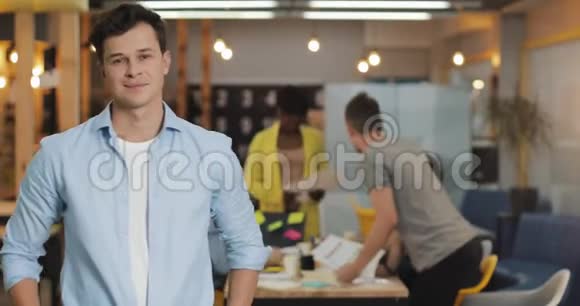 年轻微笑的成功商人站在现代办公室的肖像在模糊的背景下工作的人视频的预览图