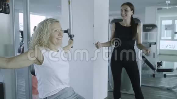 老年妇女与私人教练在健身房的模拟器上订婚女儿在健身房帮妈妈视频的预览图